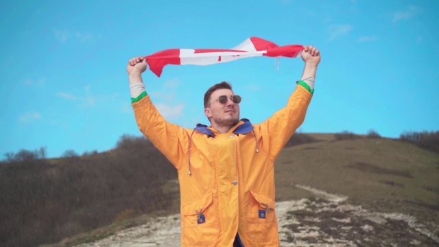 一个穿着黄色夹克，蓝色牛仔裤和眼镜的男人站在山上，双手举着加拿大国旗。视频下载