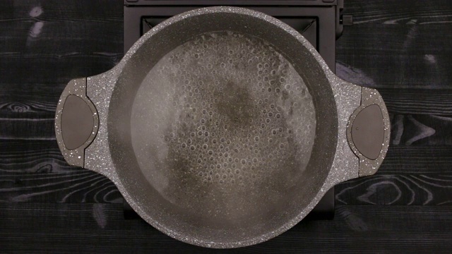 水在带有石头或大理石不粘涂层的金属锅中沸腾。平底锅放在煤气炉上，背景是一张黑色木桌。俯视图视频下载