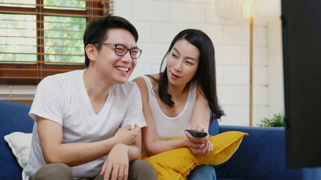 年轻的亚洲夫妇看电视与幸福在家庭客厅的沙发背景，人和家庭娱乐视频素材
