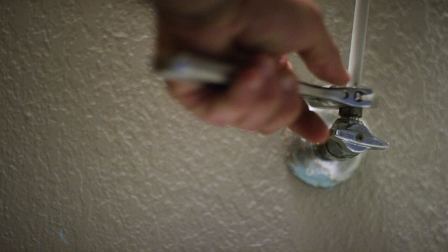 在室内家庭浴室里，一个白人修工用一个活动扳手将厕所软管连接并紧固到墙上的一个截止阀上，然后将截止阀的旋钮打开视频下载