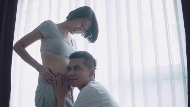 年轻的孕妇和她的丈夫抱着和听婴儿在肚子附近的窗户在家里，怀孕和家庭的概念视频素材