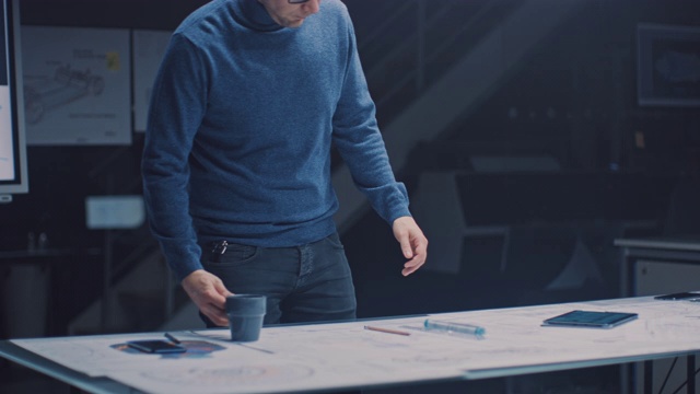 在黑暗的工业设计工程设施:男性工程师工作与蓝图铺在桌子上，使用铅笔，尺子和数字平板电脑和饮料咖啡。桌面多重绘图视频素材
