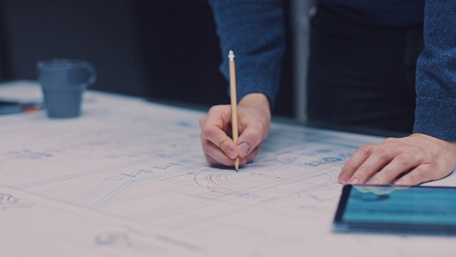 在黑暗的工业设计工程设施:男性工程师工作与蓝图铺在桌子上，使用铅笔，尺和数字平板。在桌面上的多个绘图。专注于手视频素材