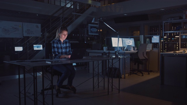 坐在办公桌前的美丽女工程师在笔记本电脑上工作。蓝图躺在桌子上。在黑暗工业设计工程设施。暖光灯视频素材