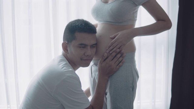 年轻的孕妇和她的丈夫抱着和听婴儿在肚子附近的窗户在家里，怀孕和家庭的概念视频素材