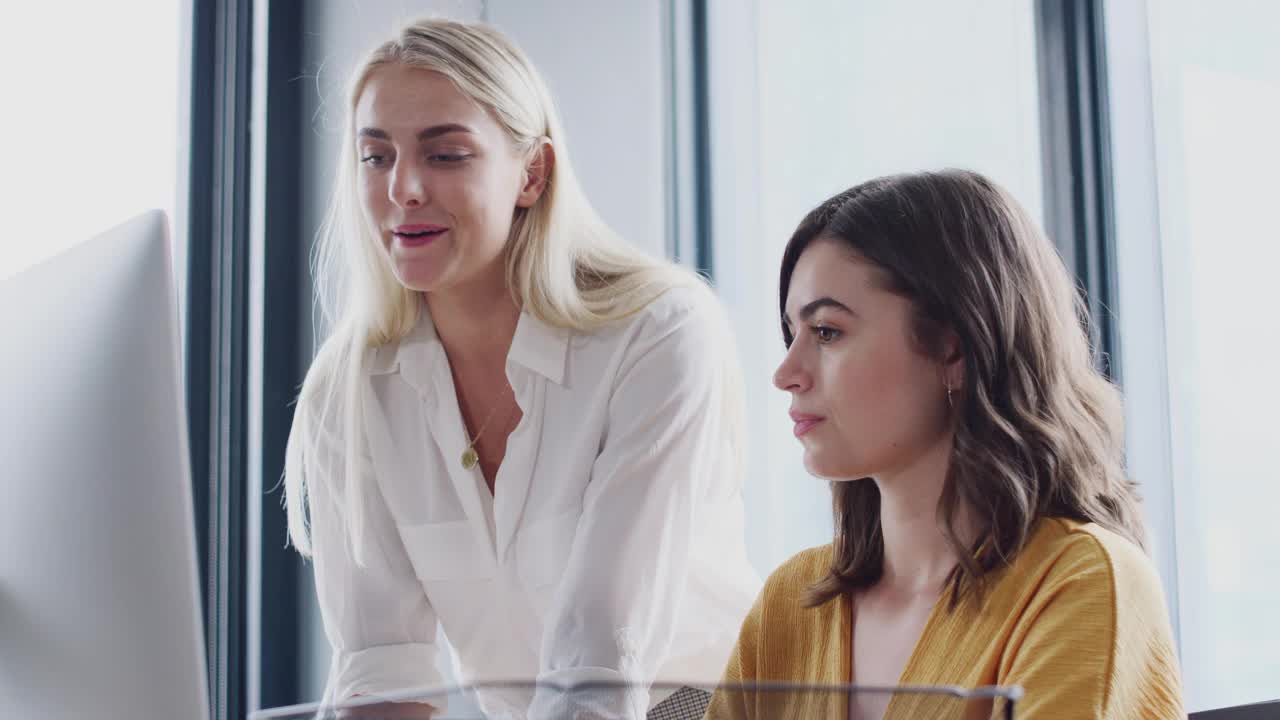 两名白人女同事在一间创意办公室的电脑屏幕前讨论视频下载