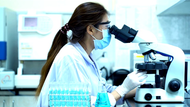 4K女科学家白色防护服看显微镜和使用移液管填充化学物质到蓝色试管在实验室做科学实验视频下载