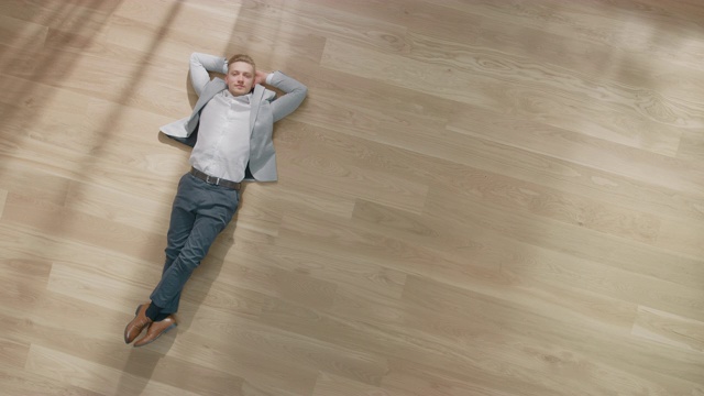 一个年轻人躺在公寓的木地板上。他穿着夹克和白衬衫。舒适的客厅与现代简约的室内和木质拼花地板。俯视图摄像机镜头。视频下载