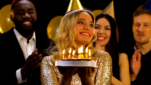 戴着生日帽的金发女人在蛋糕上吹蜡烛，朋友们在鼓掌视频素材