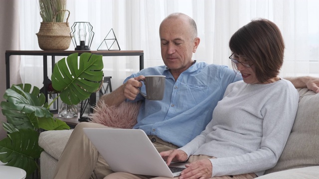 老夫妇坐在沙发上，拿着笔记本电脑和一杯咖啡视频素材