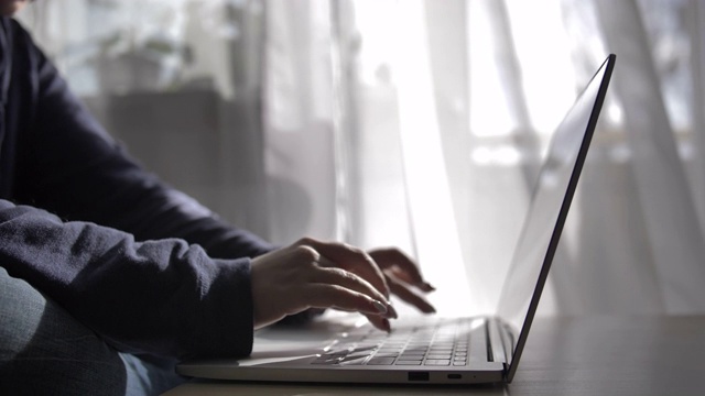 一个女人在家里的笔记本电脑上打字，背景是正在显影的窗帘视频素材