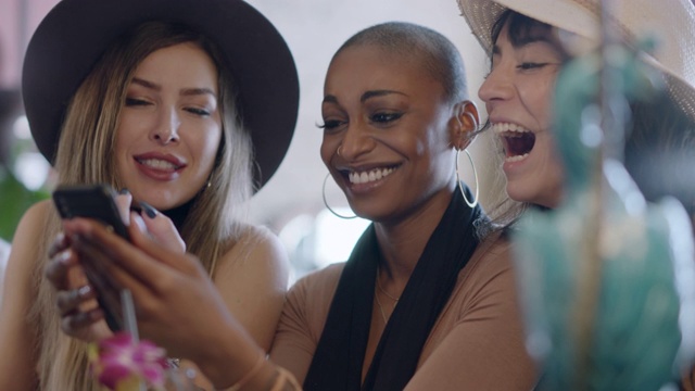 在当地酒吧的欢乐时光里，三个时髦的年轻女性一边看智能手机一边聊天、大笑。视频下载
