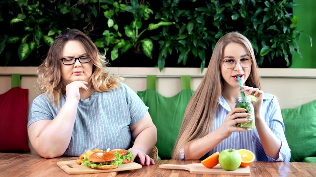 微笑苗条的女孩和悲伤的胖女人坐在咖啡馆一起快餐vs健康餐视频下载