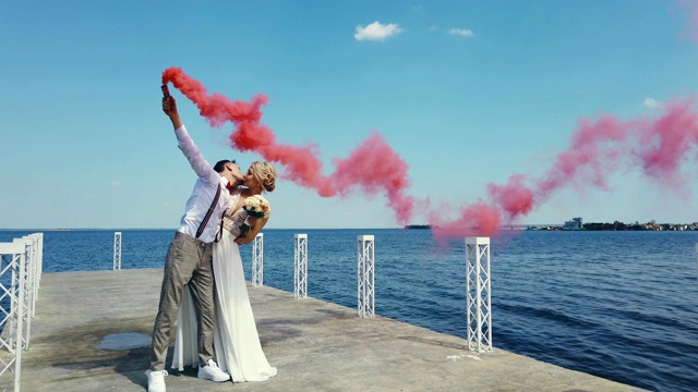 新婚夫妇用彩色烟雾的浪漫之吻，烟雾弹在手中。他们拥抱着，站在美丽的码头上，在阳光明媚的夏日里举行婚礼视频下载