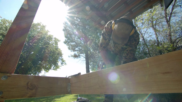 一个40多岁的白人杂工戴着帽子、太阳镜、听力保护和护腿，在一个阳光明媚的日子里，在一个居民区的户外搭建甲板时，用气动钉枪把金属角支架固定在木板上视频素材