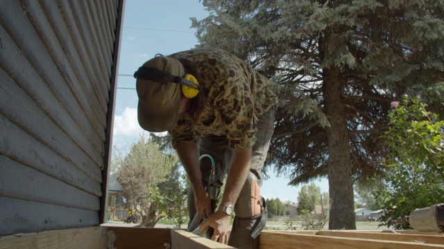 一个40多岁的白种杂工戴着一顶帽子，戴着听力保护，在一个阳光明媚的日子里，在一个居民区的户外搭建一个甲板时，用风射钉枪固定木板视频素材