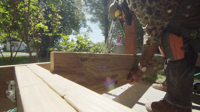 一个40多岁的白种杂工，戴着帽子、太阳镜、听力保护和护腿，在一个居民区的户外阳光下建造一个甲板时，把一块木板放在一起，然后用气动钉枪把木板固定在一起视频素材