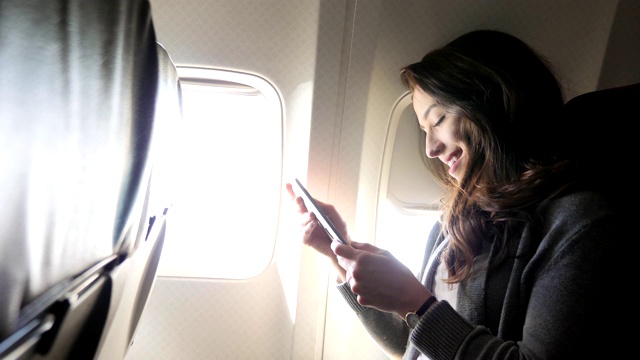 快乐的年轻女子微笑，她在飞机上使用智能手机视频素材