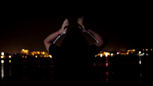 夜晚，一个戴着黑色兜帽的男人正在跳舞，从后面看过去。在水面上闪烁的城市灯光的映衬下视频素材
