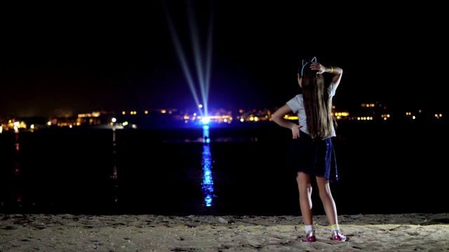 晚上，一个十几岁的女孩穿着闪亮的裙子在跳舞，从后面看。在水面上闪烁的城市灯光的映衬下视频素材