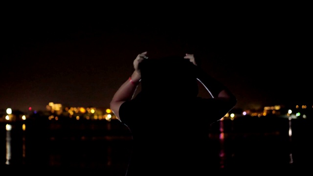 夜晚，一个戴着黑色兜帽的男人正在跳舞，从后面看过去。在水面上闪烁的城市灯光的映衬下视频素材