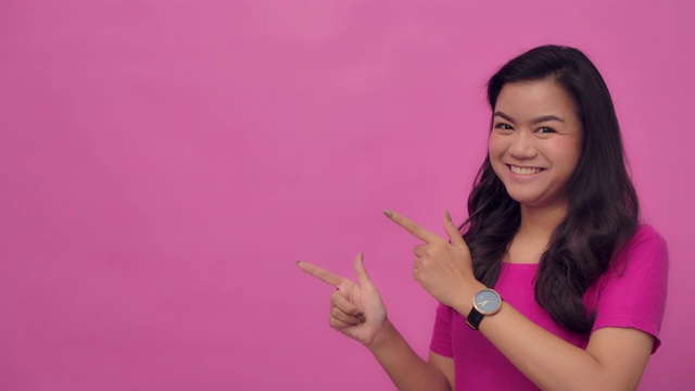 SLO MO肖像的年轻亚洲与一个幸福的微笑指向你的标志-手势在粉红色的背景视频素材