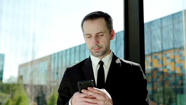 照片中，一个自信的中年商人穿着正装，站在窗边看向别处，用手机发短信视频下载
