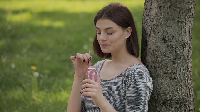 年轻积极的女人通过吸管喝新鲜的冰沙坐在夏季公园视频素材