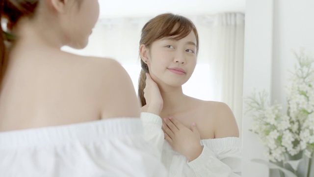 美丽的年轻亚洲女性照镜子和使用防晒乳液颈部，美丽的亚洲女孩完美的皮肤护理和健康与霜治疗和保湿，健康和保健的概念。视频素材