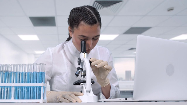 一位女性研究人员坐在一张白色的桌子前，在显微镜下观察生物样本，然后将数据写入笔记本电脑。科学实验室视频下载