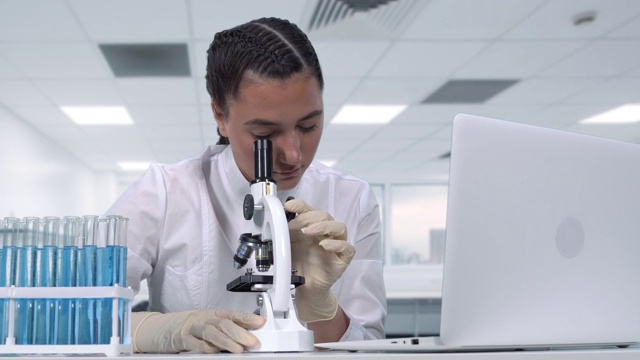 一位女性研究人员坐在一张白色的桌子前，在显微镜下观察生物样本，进行临床试验，并将数据写入笔记本电脑。科学实验室视频下载