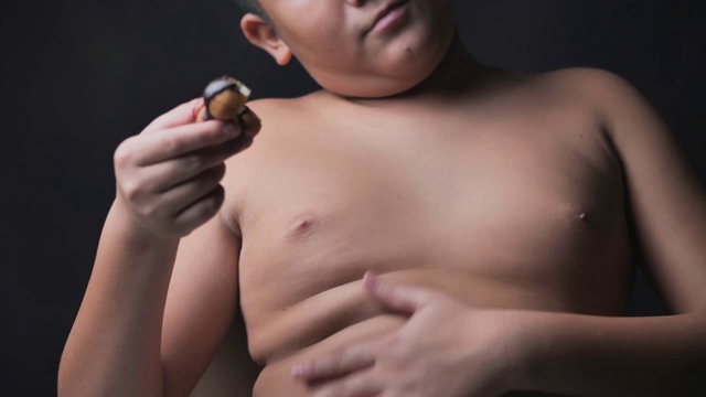 体型庞大的亚洲十岁前男孩吃甜甜圈，同时触摸他的肚子，在昏暗的光线下缓慢的动作。视频下载