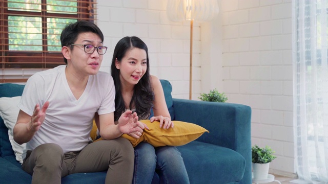 亚洲夫妇在家里的电视客厅前为足球比赛欢呼，甜蜜的夫妇在家里放松时躺在沙发上享受有趣的时刻。生活方式夫妇在家里放松的概念。视频素材