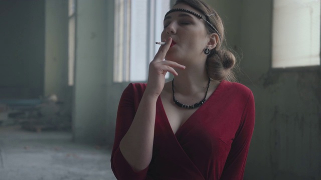 画像中优雅的女子穿着红色优雅的连衣裙坐在废弃大楼的椅子上抽烟。视频素材
