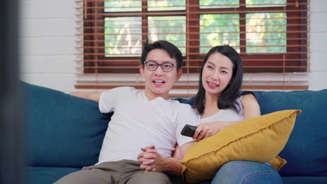亚洲夫妇在家里的客厅看电视，甜蜜的夫妇在家里放松时躺在沙发上享受爱的时刻。生活方式夫妇在家里放松的概念。视频素材