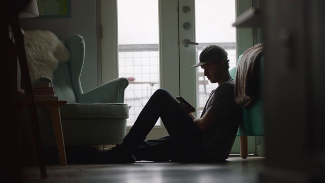 一个年轻人坐在公寓的地板上看手机。视频素材