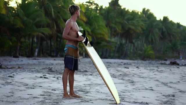 年轻健康的冲浪者看着大海。一个千禧一代在日出的海滩上，手里拿着冲浪板视频下载