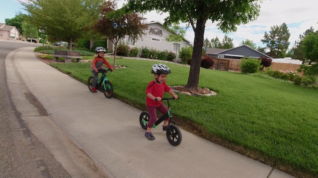 两名白人男孩(五岁和四岁)戴着自行车头盔，在多云的天空下骑着自行车穿过居民区视频素材