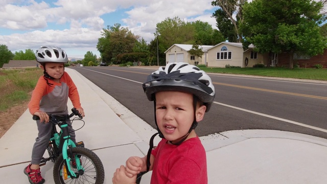 两名戴着自行车头盔的白人男孩(五岁和四岁)在多云的天空下骑着自行车穿过居民区时停下来休息视频素材