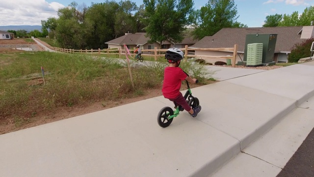 一个四岁的白人男孩戴着头盔，和他的哥哥在一个多云的天空下的住宅区骑他的平衡自行车视频素材