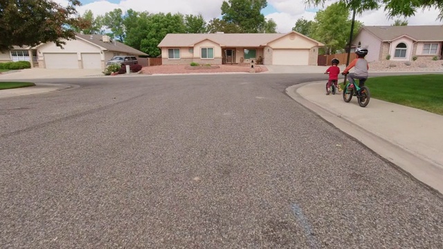 两名白人男孩(五岁和四岁)戴着自行车头盔，在多云的天空下骑着自行车穿过居民区视频素材