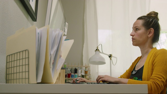一位三十多岁的白人妇女在室内家庭办公室用电脑工作，背景是缝纫材料视频素材