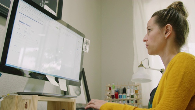 一位三十多岁的白人妇女在室内家庭办公室用电脑工作，背景是缝纫材料视频素材