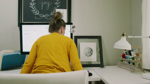 一位三十多岁的白人妇女走到她的电脑前，坐在办公椅上，打开旁边的灯，开始在室内家庭办公室的电脑上工作，背景是缝纫材料视频素材
