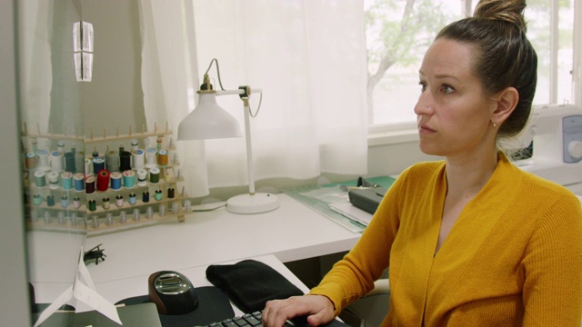 一位三十多岁的白人妇女在家里的办公室里用电脑工作，背景是一台缝纫机和一些材料视频素材