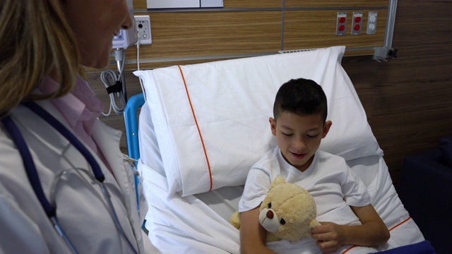 友善的女儿科医生正在检查一个住院的小男孩拥抱他的泰迪熊视频素材
