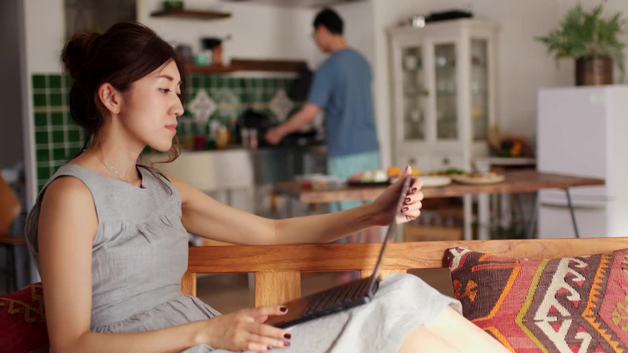 一个年轻女子使用笔记本电脑和她的男朋友谈话的广角镜头，而他正在做饭的背景视频下载