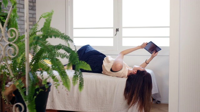 中景一名年轻女子躺在床上使用数码平板电脑视频下载