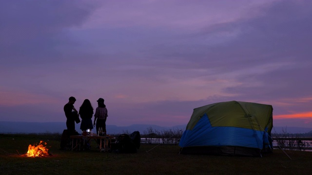 场景一群青少年亚洲人正在一起享受帐篷前的篝火和看日落附近的湖在暑假。他们是快乐的，有乐趣的假期，放松的时刻。青少年自然户外日常生活的概念视频素材