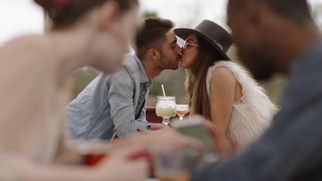 在欢乐时光，时尚的年轻夫妇斜靠在桌子上亲吻，享受着户外露台上的鸡尾酒。视频素材